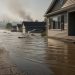 Ground-level view of a flooded street with a home partially submerged, wildfire smoke on the horizon, and heat-haze in the smoky gray sky.