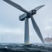 Low-angle view of a colossal floating offshore wind turbine with sweeping blades above the ocean under an overcast sky.